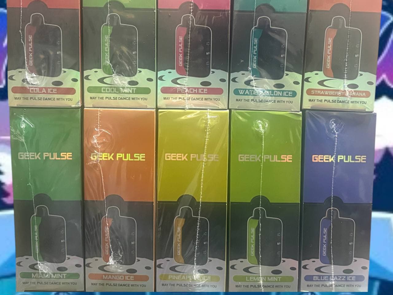 Two rows of colorful Geek Pulse disposable vape boxes in various fruit and mint flavors.